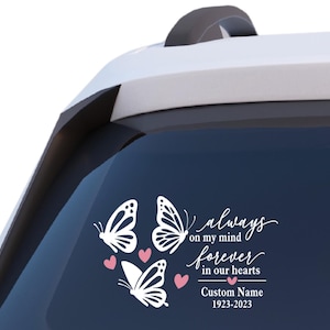 May include: A white decal featuring three butterflies and pink hearts. The text reads "always on my mind forever in our hearts" with a custom name and dates. The decal is applied to a car window.