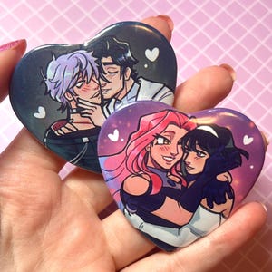 Op de afbeelding: Twee hartvormige buttons met kleurrijke illustraties van omhelzende stellen. Eén button toont twee mannen, de andere twee vrouwen. De buttons hebben een glanzende afwerking en worden in een hand gehouden.