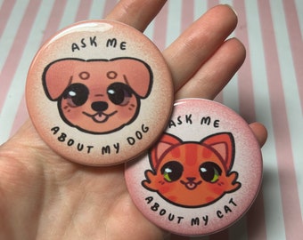 Puede incluir: Dos botones redondos rosas con ilustraciones de dibujos animados de mascotas. Uno presenta un perro con el texto "Ask Me About My Dog". El otro botón muestra un gato con el texto "Ask Me About My Cat". Ambos botones tienen un acabado brillante.