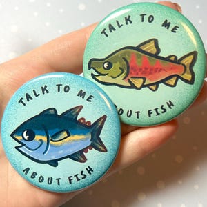 Puede incluir: Dos botones redondos con ilustraciones de peces. Un botón presenta un atún azul con aletas amarillas sobre un fondo azul claro. El otro botón tiene un pez rojo y verde sobre un fondo turquesa. Ambos botones tienen el texto "Talk to me about fish."