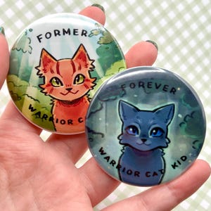 Può includere: Due bottoni rotondi con illustrazioni di gatti. Un bottone presenta un gatto arancione con la scritta "FORMER WARRIOR CAT". L'altro bottone mostra un gatto blu con la scritta "FOREVER WARRIOR CAT KID". Lo sfondo di ogni bottone è una scena di foresta.