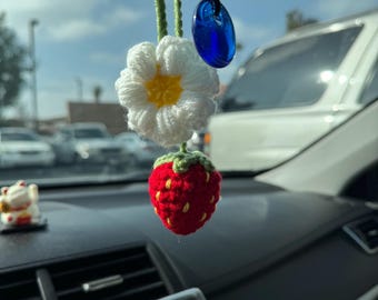 May include: A handmade car charm featuring a crocheted strawberry, daisy, and a blue glass pendant. The strawberry is red with green leaves, the daisy is white with a yellow center. The charm hangs from a green and black string.