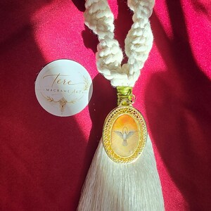 Puede incluir: Un lazo de cuerda de macramé color crema unido a un colgante ovalado dorado con la ilustración de una paloma. Una borla larga de color blanco sedoso cuelga del colgante, asegurado con hilo verde. Una pegatina circular dice Tere Macrame Art.