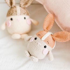May include: Two crocheted bunny plushies, one with brown ears and a beige body, the other with orange ears and a beige body. Both have pink cheeks and black eyes.