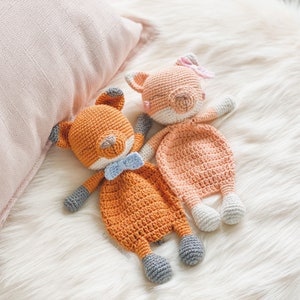Könnte beinhalten: Zwei gehäkelte Fuchs-Plüschtiere, eines orange mit einer blauen Schleife und eines rosa mit einer rosa Blume, liegen auf einer weißen, flauschigen Oberfläche.