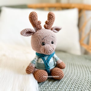 May include: A crocheted reindeer toy wearing a blue and white striped sweater and a blue scarf. The reindeer has brown antlers and a brown body. The toy is sitting on a white and green blanket.