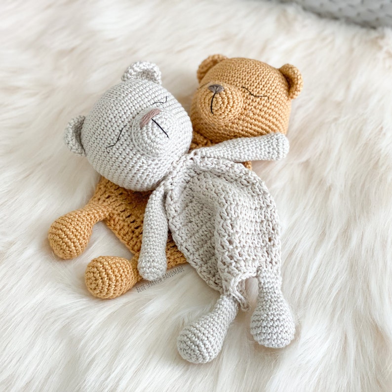 May include: Two crocheted teddy bears, one beige and one brown, are lying together on a white fluffy surface. The beige bear is on top of the brown bear, with its head resting on the brown bear's back. Both bears have closed eyes and are made with a soft, textured yarn.