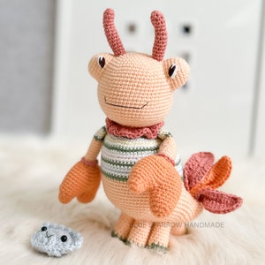 May include: A crocheted plush toy crab with pink and orange accents. The crab is wearing a striped shirt and has a friendly expression. The toy is standing on a light brown surface next to a small gray crocheted clam.