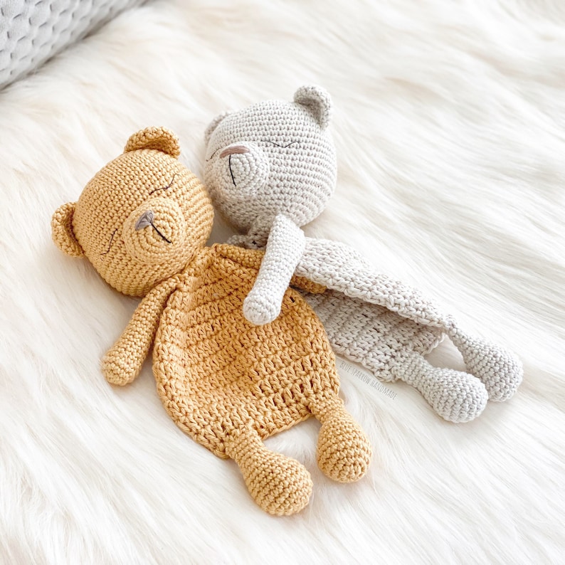 May include: Two crocheted teddy bear security blankets, one in beige and one in mustard yellow. Both bears have closed eyes and are lying on a white fluffy surface.