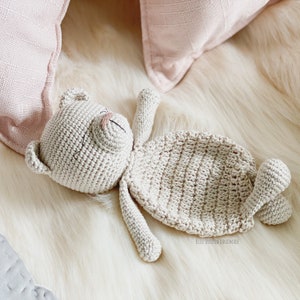 May include: A crocheted beige teddy bear lying on a white fluffy surface. The bear has a stitched-on nose and eyes.