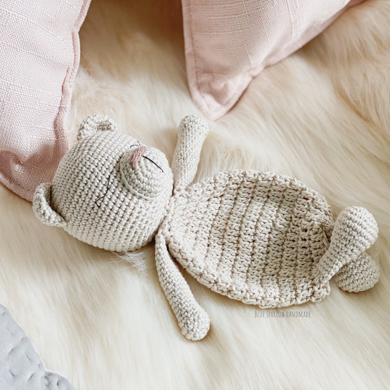 May include: A crocheted beige teddy bear lying on a white fluffy surface. The bear has a stitched-on nose and eyes.