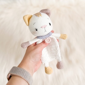 May include: A crocheted cat toy with a white body, pink and yellow accents, and a purple collar. The cat is holding a white crocheted blanket with a pink flower.