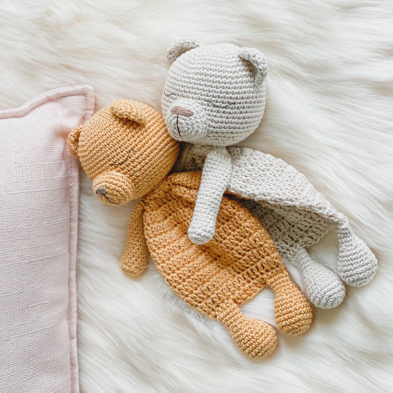 May include: Two crocheted stuffed bears, one beige and one orange, are lying on a white fluffy surface. The beige bear is on top of the orange bear, with its head resting on the orange bear's back. Both bears have closed eyes and are sleeping.