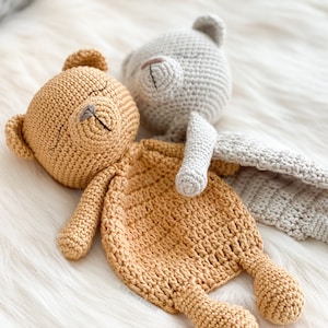 May include: Two crocheted teddy bears, one brown and one gray, are lying on a white fluffy surface. The brown bear is lying on its back with its arms outstretched, while the gray bear is lying on its side with its head resting on the brown bear's arm. The bears are both wearing a crocheted blanket.