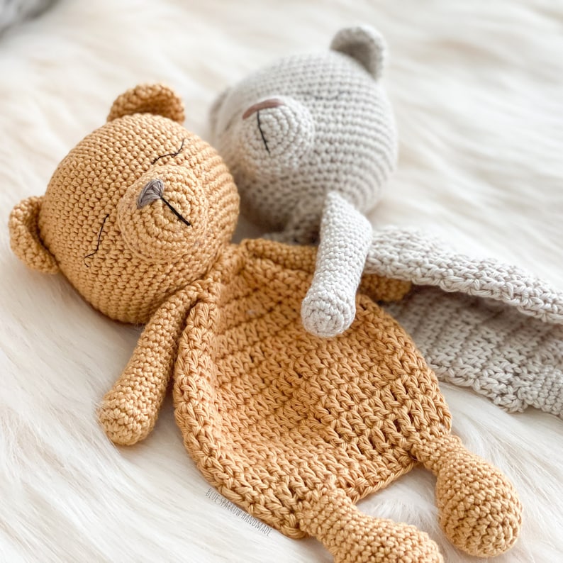 May include: Two crocheted teddy bears, one brown and one gray, are lying on a white fluffy surface. The brown bear is lying on its back with its arms outstretched, while the gray bear is lying on its side with its head resting on the brown bear's arm. The bears are both wearing a crocheted blanket.