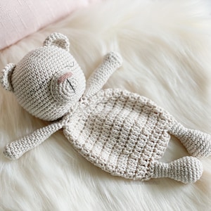 May include: A crocheted beige teddy bear with closed eyes and a stitched nose lies on a white fluffy surface.