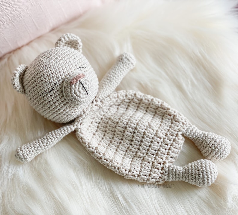 May include: A crocheted beige teddy bear with closed eyes and a stitched nose lies on a white fluffy surface.