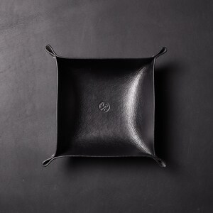 May include: A black leather catchall tray with a small circular logo in the center.