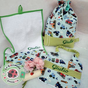 Può includere: Un set di articoli per bambini, tra cui un asciugamano, una borsa con coulisse, un bavaglino e un libro tranquillo. Gli articoli presentano un tessuto azzurro con illustrazioni di veicoli colorati e il nome "Giulio" ricamato in verde. È presente anche un piccolo bouquet di fiori rosa.