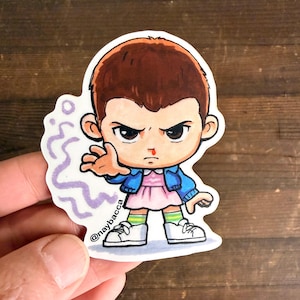 Puede incluir: Una pegatina con una ilustración de dibujos animados de un personaje con cabello castaño, que lleva un vestido rosa, una chaqueta azul y zapatillas blancas. El personaje tiene la nariz ensangrentada y una expresión enfadada. La pegatina tiene el texto "@naybacca".