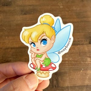 Peut inclure: Autocollant représentant une fée de dessin animé aux cheveux blonds, aux yeux bleus et aux ailes bleu clair. Elle est assise sur un champignon rouge à pois blancs. Le sticker porte le texte "@naybacca".