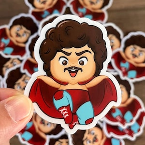 May include: A sticker featuring a cartoon luchador character with brown hair, a mustache, and a red cape. The luchador wears red trunks and boots with blue accents. The sticker has a white border and is held in a hand.