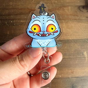 Pode incluir: Um porta-crachá de acrílico transparente com um gato de desenho animado azul-claro com grandes olhos amarelos, dentes afiados e um pequeno pássaro preto empoleirado na cabeça. O gato está na água com ondulações e o texto naybacca está impresso.