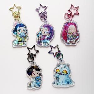 Op de afbeelding: Vijf acryl sleutelhangers met cartoonkarakterontwerpen. Elke sleutelhanger heeft een ander karakter, met stervormige sluitingen in goud, zilver en roze. De karakters zijn onder meer een meisje met blauw haar, een vrouw met paars haar en een kat.