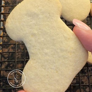 May include: A light-colored, gluten-free cookie in the shape of a stocking, held by a hand with a painted nail. The cookie is on a black wire rack. The cookie has the text "Gluten Free Baked by Dee" in a circular logo.