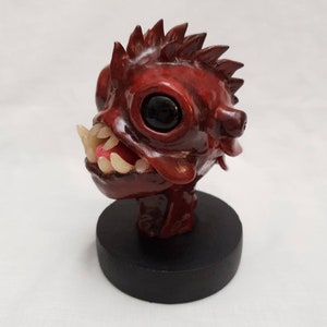 May include: A red, hand-painted, ceramic sculpture of a creature with a large mouth, sharp teeth, and one large eye. The sculpture is mounted on a black base.