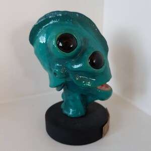 May include: A teal green alien bust with large black eyes and an open mouth. The bust is on a black circular base.