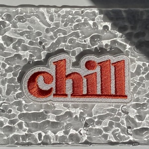 Könnte beinhalten: Ein weißer Patch mit dem Wort "chill" in orangefarbenen Buchstaben auf einem strukturierten, transparenten Hintergrund.