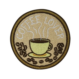 Può includere: Una toppa ricamata marrone con il testo "Coffee Lover" in bianco. Un'immagine di cartone animato di una tazza di caffè fumante con un manico marrone e un piattino è al centro della toppa. I chicchi di caffè marroni sono intorno alla tazza e al piattino.