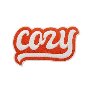 Könnte beinhalten: Ein gestickter Aufnäher mit dem Wort "cozy" in weißer Schrift. Das Wort ist orange umrandet, mit einem weißen Rand. Der Aufnäher hat eine geschwungene Form und ist vor einem weißen Hintergrund platziert.