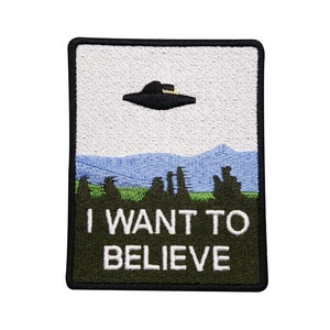 Könnte beinhalten: Ein gestickter Patch in Grün und Schwarz mit einem schwarzen UFO, das über einem grünen Feld und blauen Bergen fliegt. Der Text "I WANT TO BELIEVE" ist in Weiß auf dem Patch geschrieben.