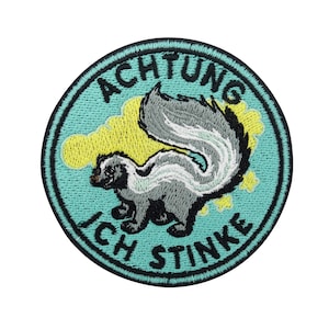 Könnte beinhalten: Ein runder, türkisblauer Patch mit schwarzen Nähten. Der Patch zeigt eine Zeichentrick-Stinktier mit einem weißen Streifen und dem Text "ACHTUNG ICH STINKE" in Schwarz.