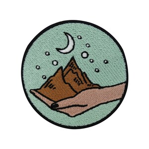 Könnte beinhalten: Ein grüner gestickter Aufnäher mit einer braunen Bergkette und einem Halbmond am Himmel. Die Bergkette wird in zwei Händen gehalten.