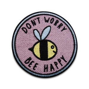 Może przedstawiać: Okrągła, haftowana naszywka z jasnofioletowym tłem i czarną obwódką. Naszywka przedstawia żółto-czarną pszczołę z napisem "DON'T WORRY BEE HAPPY" czarnymi literami. Pszczoła ma jasnoniebieskie skrzydła.