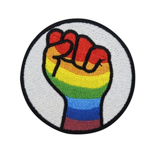 Op de afbeelding: Een ronde geborduurde patch met een regenboogvuist die in de lucht geheven is. De vuist is zwart omlijnd en de achtergrond is wit.