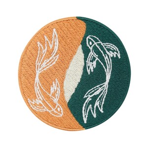 May include: A round embroidered patch featuring two koi fish swimming in opposite directions. The fish are white and the background is split into two sections: one orange and one green.