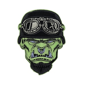 Könnte beinhalten: Gestickter Aufnäher mit einem grünen, ork-ähnlichen Gesicht, das einen schwarzen Motorradhelm mit Brille trägt. Der Ork hat einen schwarzen Bart, blaue Augen und einen Nasenring. Der Aufnäher befindet sich auf weißem Hintergrund.