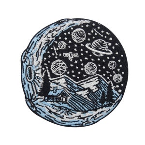 May include: A black and white embroidered patch featuring a crescent moon, stars, planets, and a small house in a mountain landscape. The patch is circular and has a blue outline.