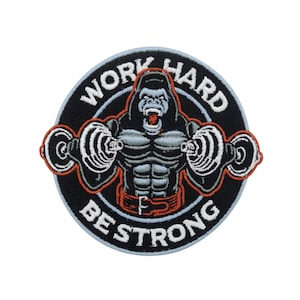 Può includere: Una toppa ricamata in bianco e nero con un gorilla che solleva pesi. La toppa ha il testo "Work Hard Be Strong" attorno al bordo.