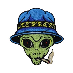 Op de afbeelding: Een geborduurde patch van een groene alien die een blauwe hoed draagt met een marihuanabladontwerp en een joint rookt.