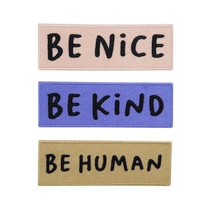 Könnte beinhalten: Drei rechteckige Aufnäher mit schwarzem Text auf unterschiedlich farbigen Hintergründen. Der obere Aufnäher ist hellrosa mit "BE NICE". Der mittlere Aufnäher ist blau mit "BE KIND". Der untere Aufnäher ist beige mit "BE HUMAN".