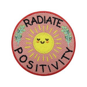 May include: An embroidered patch in pink and red with a yellow sun with a smiling face and the text "RADIATE POSITIVITY" in black.