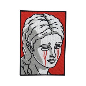May include: An embroidered patch featuring a woman's face with red tears. The background is red.