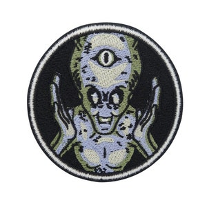 Op de afbeelding: Gestikte patch met een groene alien met één oog en een brede glimlach, met uitgestrekte armen, tegen een zwarte achtergrond.