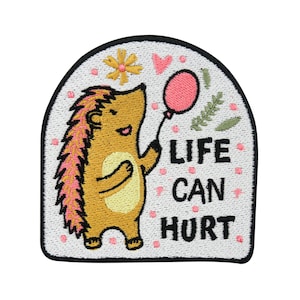 Könnte beinhalten: Gestickter Bügel-Patch mit einem Comic-Igel, der einen rosa Ballon hält, mit dem Text "Life can hurt" in Weiß.