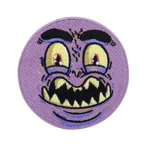 Puede incluir: Parche bordado morado con una cara de monstruo de dibujos animados con ojos amarillos, una amplia sonrisa de dientes y el ceño fruncido.